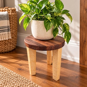 May include: A small round wooden plant stand with a dark brown circular top and three light-colored tapered legs. A white speckled ceramic pot sits on the stand, containing a vibrant green and yellow variegated pothos houseplant.