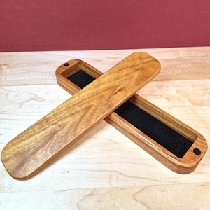 May include: A long, rectangular wooden box with a matching lid placed diagonally across it. The box features a natural light brown wood grain finish and a deep black velvet interior lining. Small black circular magnets are visible at the ends.