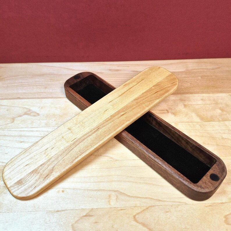 May include: A long, rectangular wooden box with a dark brown base and a light tan wooden lid placed diagonally across it. The interior of the box is lined with black velvet, and the lid features rounded edges on both ends of the piece.