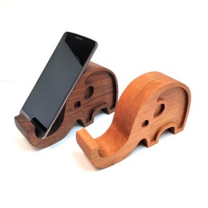 Peut inclure: Deux supports de téléphone en bois en forme d'éléphant, l'un brun foncé et l'autre brun clair. Les supports sont en forme d'éléphants avec leurs trompes levées pour tenir un téléphone.