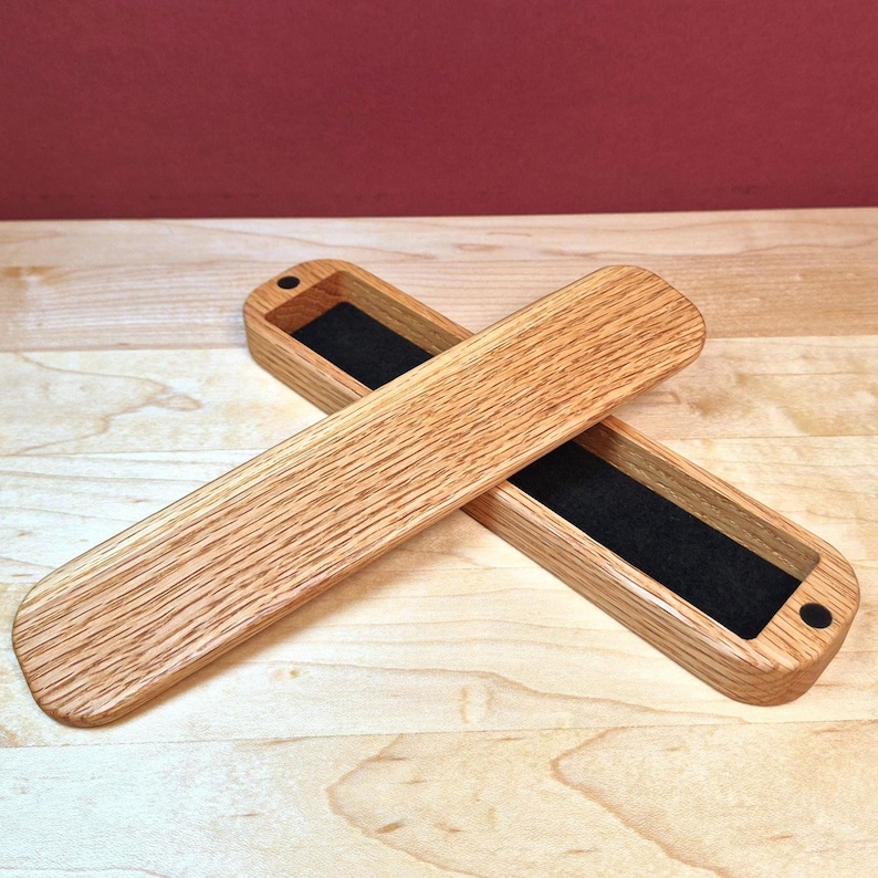 May include: A long, rectangular wooden box with a matching lid, both featuring a natural light brown wood grain finish. The interior of the box is lined with black felt, and the lid is secured by small circular magnets at each end.