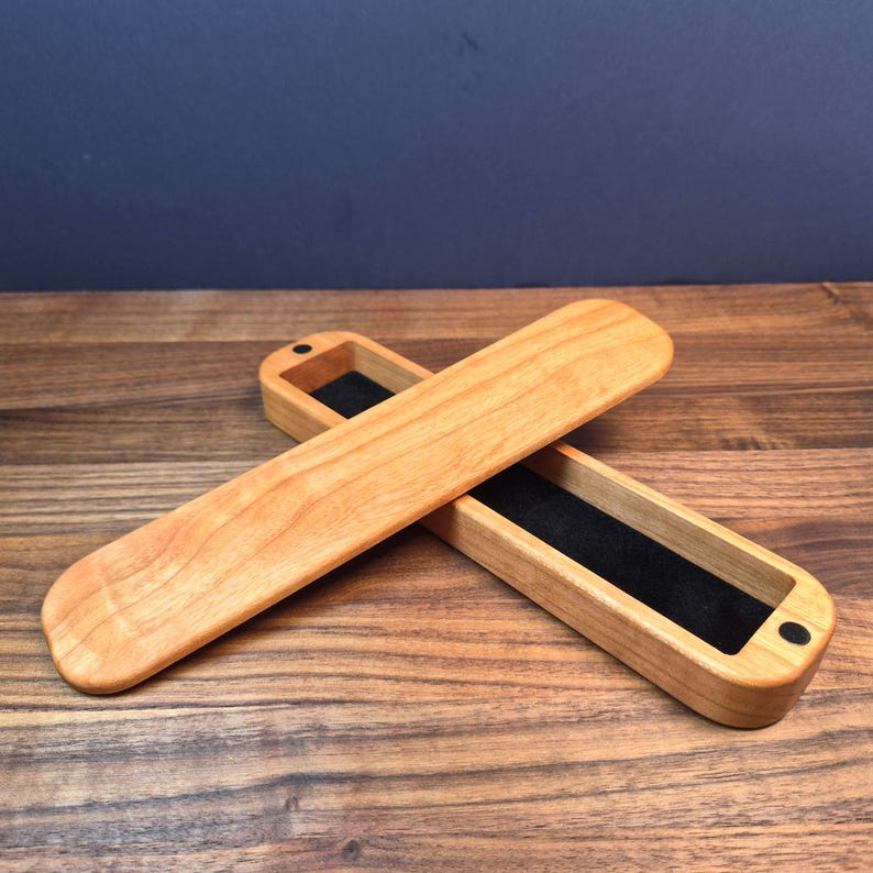 May include: A long, rectangular wooden box with a smooth, light-toned finish and rounded ends. The box is open, revealing a black velvet-lined interior. A matching wooden lid is positioned diagonally across the open base, featuring small black circular magnets at each end for closure.