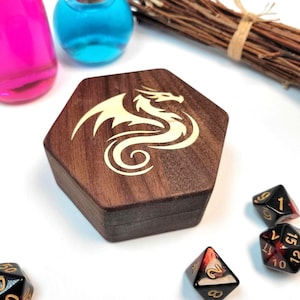 May include: A dark brown hexagonal wooden box featuring a cream-coloured dragon design on the lid. Several dice are scattered around the box, along with two glass bottles and a bundle of twigs.