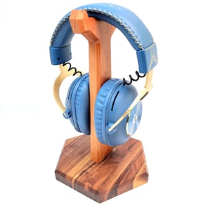 May include: Blue headphones with gold detailing sit on a wooden headphone stand. The headphones feature a stitched headband and circular ear cups. The stand is made of light brown wood and has a hexagonal base of darker wood.
