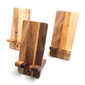 May include: Three wooden phone stands in varying shades of brown and tan. Each stand has a rectangular base and a vertical back with a slot to hold a phone. The wood grains and colors vary, showcasing natural wood patterns.