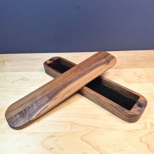 May include: A long, rectangular wooden box with rounded ends, featuring a dark brown wood grain finish. The box is open, revealing a black velvet-lined interior. A matching wooden lid is placed diagonally across the open base of the box.