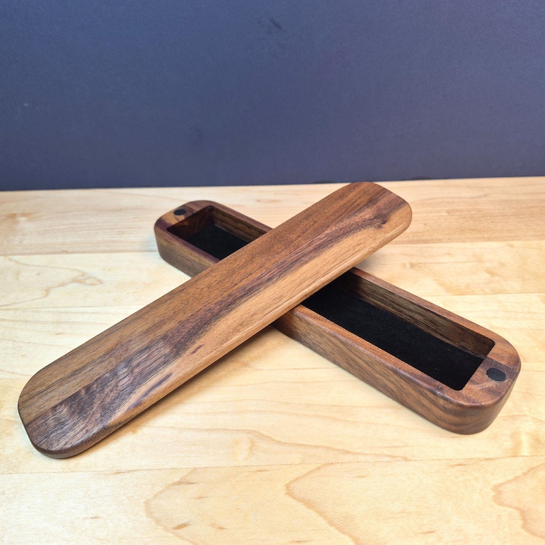 May include: A long, rectangular wooden box with rounded ends, featuring a dark brown wood grain finish. The box is open, revealing a black velvet-lined interior. A matching wooden lid is placed diagonally across the open base of the box.