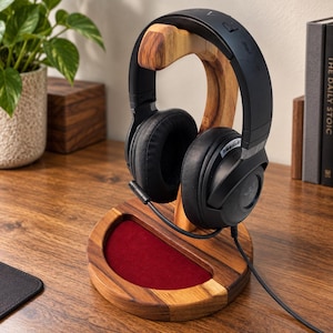 May include: A wooden headphone stand with a curved support arm and a circular base featuring a recessed red velvet tray. Black over-ear headphones with a wired connection rest on the stand, showcasing a textured ear cup and a flexible microphone.