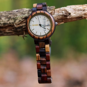 Puede incluir: Un reloj de pulsera de madera con una esfera blanca y agujas negras. El reloj tiene una correa de madera marrón, negra y roja. La esfera del reloj tiene la palabra "Joycoast" impresa en ella y los días de la semana están listados alrededor del borde.