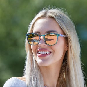 May include: A woman wearing sunglasses with a green and blue floral pattern. The sunglasses have a brown lens and a thin black frame.