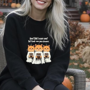 Puede incluir: Una sudadera negra con el texto "Boo! Did I scare you? No? Good - now pass Creamer." y un gráfico de tres gatos disfrazados de fantasmas sosteniendo tazas de café. La escena está ambientada al aire libre con decoraciones de Halloween.
