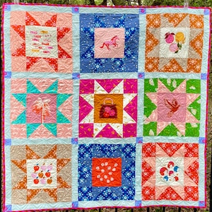 May include: A patchwork quilt with nine squares, each featuring a different design. The squares are arranged in a 3x3 grid. The designs include a star with a unicorn, a star with fruit, a star with a purse, a star with a bird, a star with a heart, a star with a heart, a star with a heart, a star with a heart, and a star with a heart.