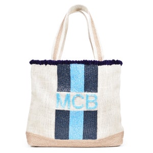 May include: A white canvas tote bag with blue and black stripes and the letters "MCB" in blue glitter. The bag has a navy blue pom-pom trim and natural jute handles.