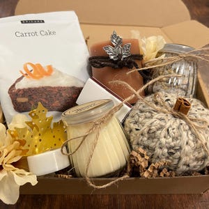 May include: A gift basket filled with fall-themed items, including a bag of carrot cake mix, a crocheted pumpkin, a candle, a bar of soap, and a jar of bath salts. The basket is lined with brown paper and tied with twine.