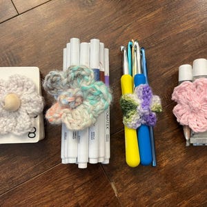 May include: An assortment of craft supplies, including white markers, crochet hooks, and tubes of glue, each adorned with handmade crochet flower embellishments. The flowers are in various colors, including pink, blue, and green, and are arranged on a wooden surface.