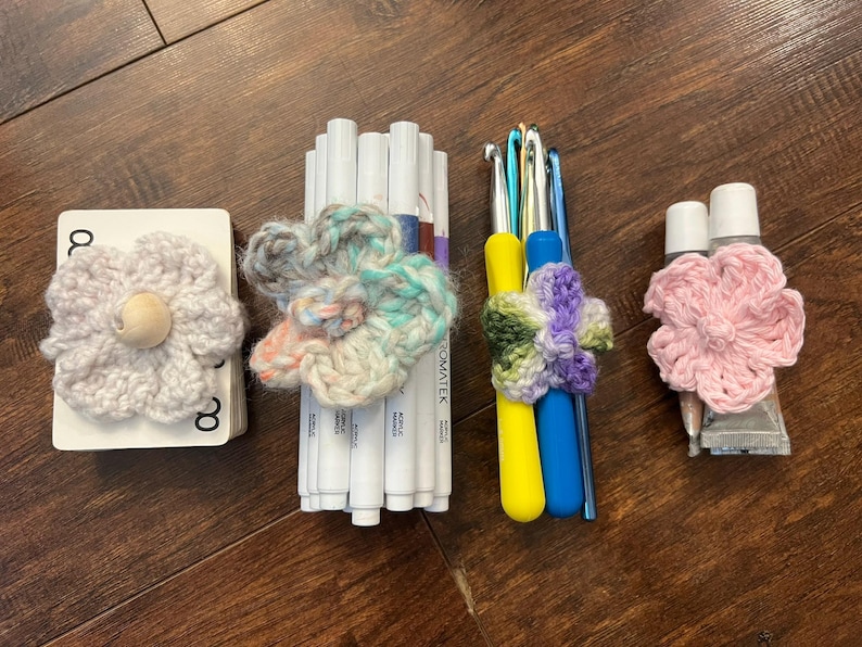 May include: An assortment of craft supplies, including white markers, crochet hooks, and tubes of glue, each adorned with handmade crochet flower embellishments. The flowers are in various colors, including pink, blue, and green, and are arranged on a wooden surface.