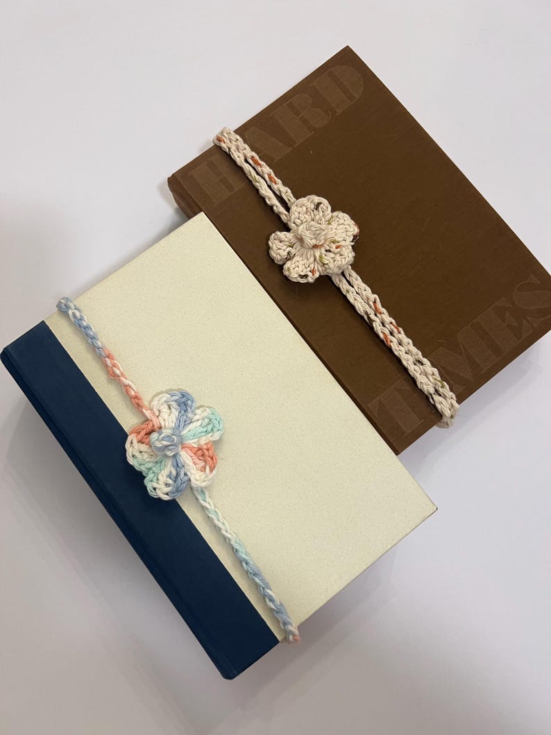 May include: Two books with decorative crochet flower bookmarks. One book is brown with embossed text, the other is cream with a navy blue spine. The bookmarks feature a flower design in cream and pastel colors.