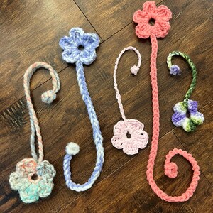 May include: A collection of handmade crochet flower bookmarks in various colors. The bookmarks feature flower designs in shades of pink, blue, green, and white. Each bookmark has a long, braided stem with a loop or a small ball at the end.
