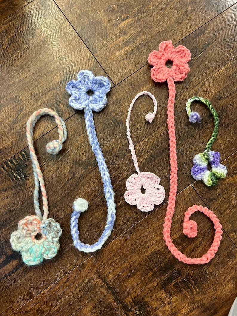 May include: A collection of handmade crochet flower bookmarks in various colors. The bookmarks feature flower designs in shades of pink, blue, green, and white. Each bookmark has a long, braided stem with a loop or a small ball at the end.