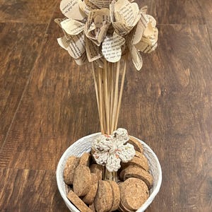 May include: A decorative arrangement featuring paper flowers made from book pages, set in a white woven bowl filled with corks. A crocheted flower accents the arrangement. The flowers are on wooden dowels.