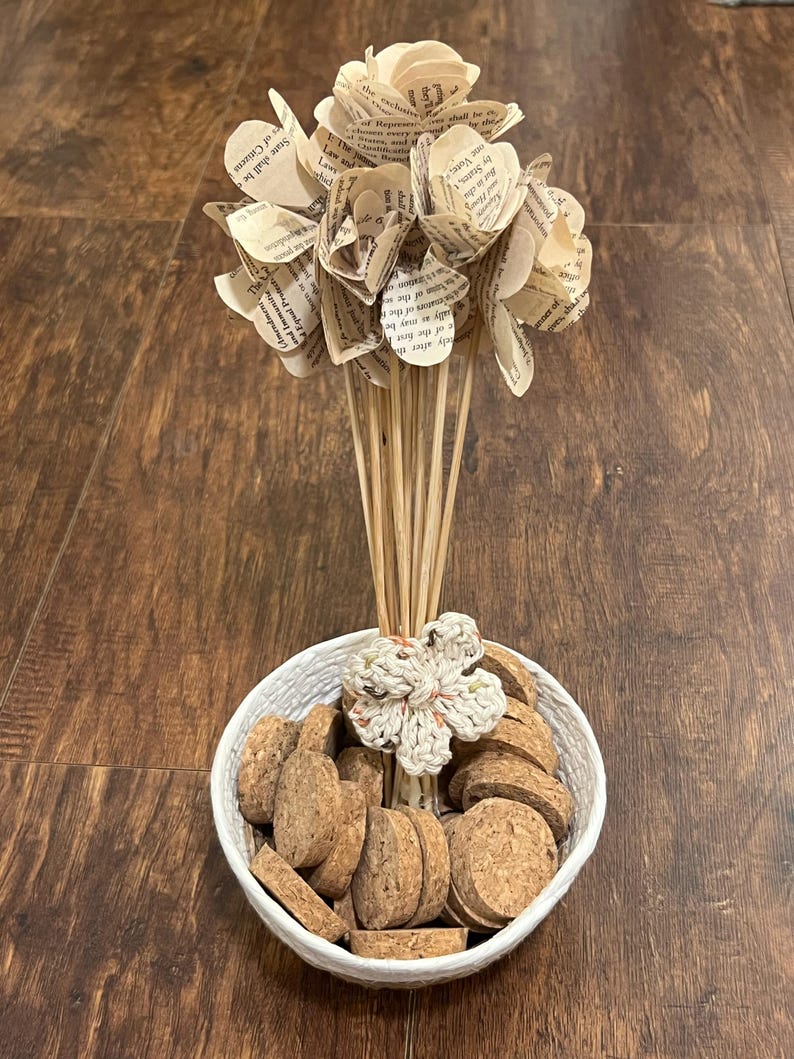 May include: A decorative arrangement featuring paper flowers made from book pages, set in a white woven bowl filled with corks. A crocheted flower accents the arrangement. The flowers are on wooden dowels.
