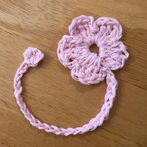 May include: A handmade, light pink crochet flower with a matching stem and bud. The flower has five petals and a circular center. The stem and bud are connected to the flower, creating a decorative element.