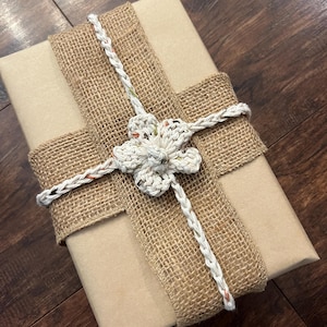 Op de afbeelding: Een ingepakt cadeau met een beige papieren omslag. Het is versierd met een breed jute lint en een gehaakte bloem. De bloem en het lint zijn wit met kleurrijke accenten. Het cadeau ligt op een donkere houten ondergrond.
