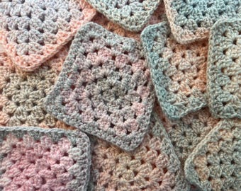 May include: A collection of square crochet motifs featuring a granny square pattern. The yarn displays a variegated color palette of soft pastel pink, muted peach, and light grey tones, creating a textured, interconnected fabric surface.
