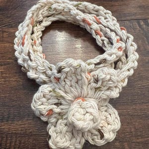 May include: Cream-colored, hand-crocheted scarf with a large flower accent. The scarf is made of thick yarn with flecks of orange, green, and brown. The flower is centered and has multiple petals. The scarf is displayed on a dark wood surface.