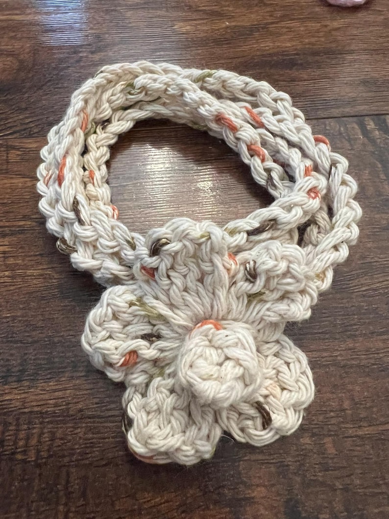 May include: Cream-colored, hand-crocheted scarf with a large flower accent. The scarf is made of thick yarn with flecks of orange, green, and brown. The flower is centered and has multiple petals. The scarf is displayed on a dark wood surface.