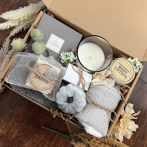 May include: A gift box filled with self-care items, including a gray towel, a candle, a bar of soap, a small bottle of lotion, a daily planner, and a decorative pumpkin.