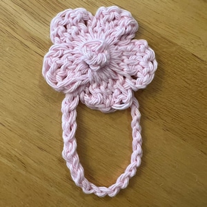 May include: A handmade, light pink crochet flower with a loop handle. The flower has a detailed petal design and a central knot. The loop is made of braided yarn, suitable for a towel or decorative item.