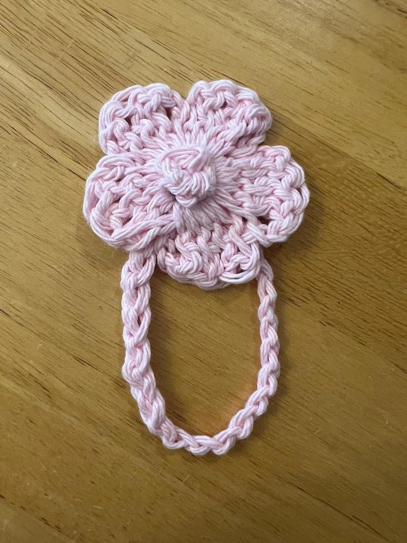 May include: A handmade, light pink crochet flower with a loop handle. The flower has a detailed petal design and a central knot. The loop is made of braided yarn, suitable for a towel or decorative item.