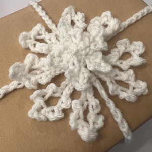 Puede incluir: Un copo de nieve blanco tejido a ganchillo adorna una caja de regalo marrón. El copo de nieve presenta un diseño intrincado con delicados patrones calados. Un cordón blanco tejido a ganchillo a juego envuelve la caja, realzando la presentación festiva. El tema general es el envoltorio de regalos navideños.