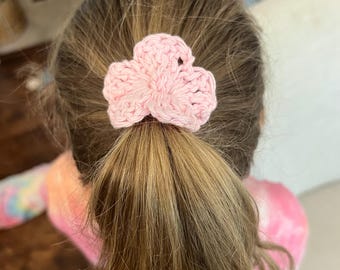 May include: A light pink, crocheted hair tie in the shape of a flower is secured in a ponytail. The hair tie is made of a soft, textured yarn. The hair is a light brown color.