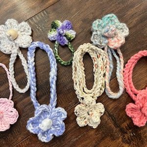 May include: A collection of handmade crochet flower accessories in various colors, including pink, blue, and white. Each flower is attached to a braided cord, creating a unique and decorative item.