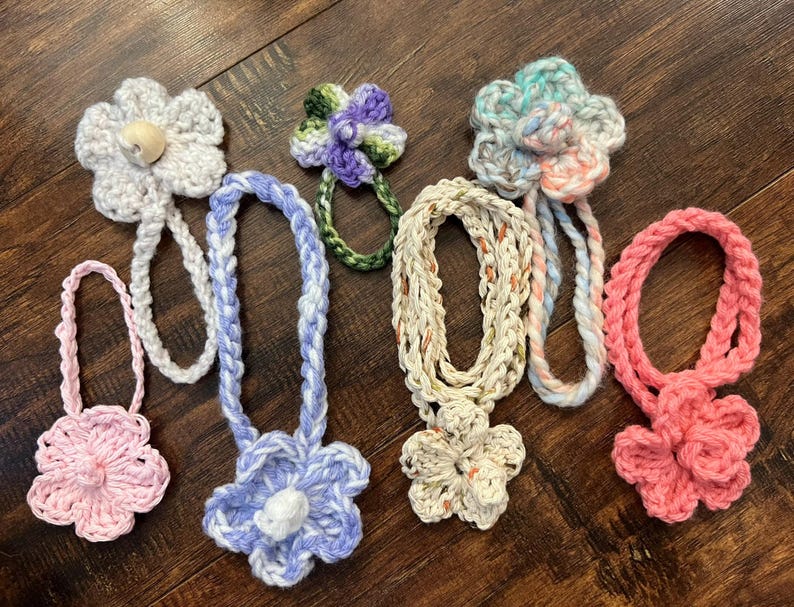 May include: A collection of handmade crochet flower accessories in various colors, including pink, blue, and white. Each flower is attached to a braided cord, creating a unique and decorative item.