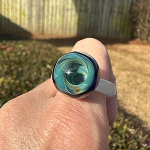 May include: A handcrafted glass ring with a swirling pattern of teal, blue, and gold colors. The ring has a white band and is worn on a finger. The glass design resembles a miniature galaxy or ocean vortex.