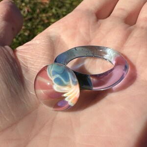 May include: A clear glass ring and a glass orb with a colorful design of pink, blue, and white swirls. The ring is a gradient of blue and pink. The orb has a similar color scheme and design. The items are displayed on a hand.
