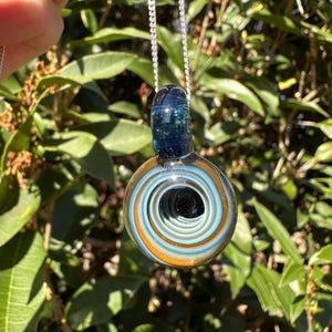 May include: A glass pendant necklace with a spiral design in shades of blue and orange. The pendant is attached to a silver chain. The top of the pendant is a dark blue color. The background is green foliage.