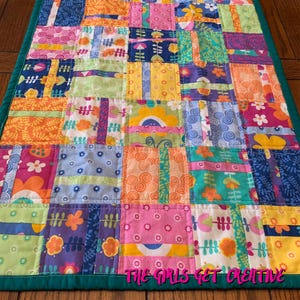 May include: A rectangular patchwork quilt featuring a grid of colorful fabric squares with various floral, swirl, and geometric patterns in shades of pink, orange, yellow, blue, and green, finished with a solid dark teal border and stitched edges.