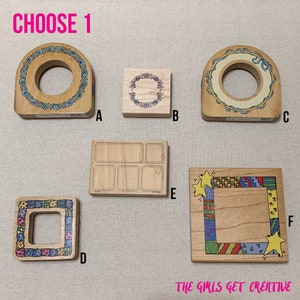 May include: Wooden stamps with various designs, including a floral wreath, a square frame with colorful patterns, and a rectangular stamp with multiple boxes. The text "CHOOSE 1" is at the top, and "THE GIRLS GET CREATIVE" is at the bottom.