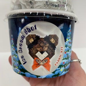 May include: A cylindrical container with a clear plastic lid containing a fabric kit. The label features a dark blue winter scene with trees and a central circular graphic of a brown patchwork bear with an orange bow and snowshoe charm.