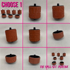 May include: Nine different brown rubber stamps with various designs, including words, patterns, and images. The stamps are arranged in a grid on a beige surface. The text "CHOOSE 1" is at the top of the image. The text "THE GIRLS GET CREATIVE" is at the bottom of the image.
