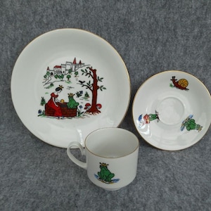 Può includere: Un servizio da tè per bambini bianco con finiture dorate. Il set include un piatto, un piattino e una tazza, ciascuno decorato con illustrazioni colorate di una principessa, rane e una lumaca. Il piatto presenta una scena di castello.