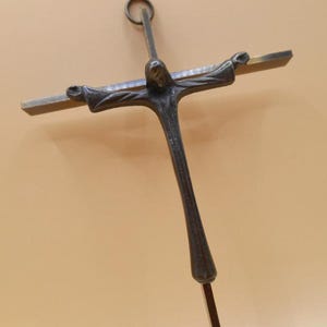 Könnte beinhalten: Ein dunkles Metallkruzifix mit einer detaillierten Figur Jesu. Das Kreuz hat einen vertikalen Pfosten und einen horizontalen Balken, mit einem Ring oben zum Aufhängen. Die Arme der Figur sind ausgestreckt.