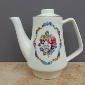 May include: Cream-colored ceramic coffee pot with a floral design. The oval floral arrangement features a pink rose, white flower, and various other colorful blooms. The pot has a gold trim and a handle.