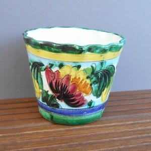 Puede incluir: Una maceta de cerámica con borde festoneado, con un diseño floral. La maceta tiene un interior blanco, con un exterior azul claro adornado con flores rojas, amarillas y verdes. Bandas verdes y amarillas rodean la parte superior y una banda azul acentúa la base.