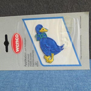 Könnte beinhalten: Ein blaues Enten-Aufbügler mit gelben Füßen und Schnabel sowie einer grünen Schleife. Der Aufbügler befindet sich auf einer grauen Rückseite mit dem Markennamen "wenco" und Text auf Deutsch, Französisch und Englisch.