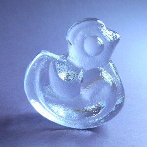 Puede incluir: Figura de pato de cristal transparente con superficie texturizada. El pato tiene un cuerpo redondeado, un pico pequeño y un ojo circular. La figura está sobre una superficie azul claro.